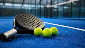 Tre padelbolde ved et perforeret padelbat på blå kunstgræs